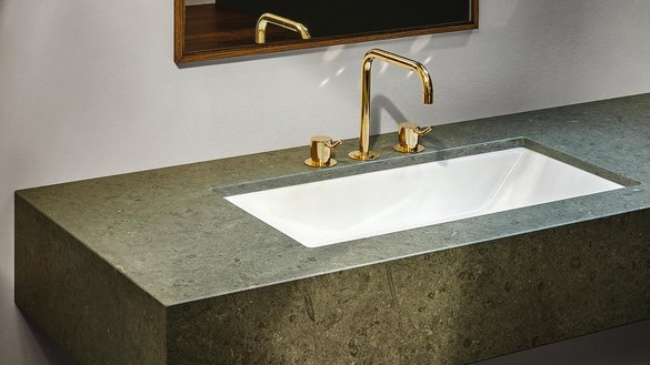 Ein elegantes Waschbecken aus weißem Porzellan, umgeben von einer stilvollen, grauen Steinplatte und goldenen Wasserhähnen.