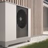 Moderne Wärmepumpe neben einem stilvollen Holzhaus Eine moderne Wärmepumpe steht neben einem Holzpaneel, umgeben von grünem Gras und einem gepflasterten Weg.